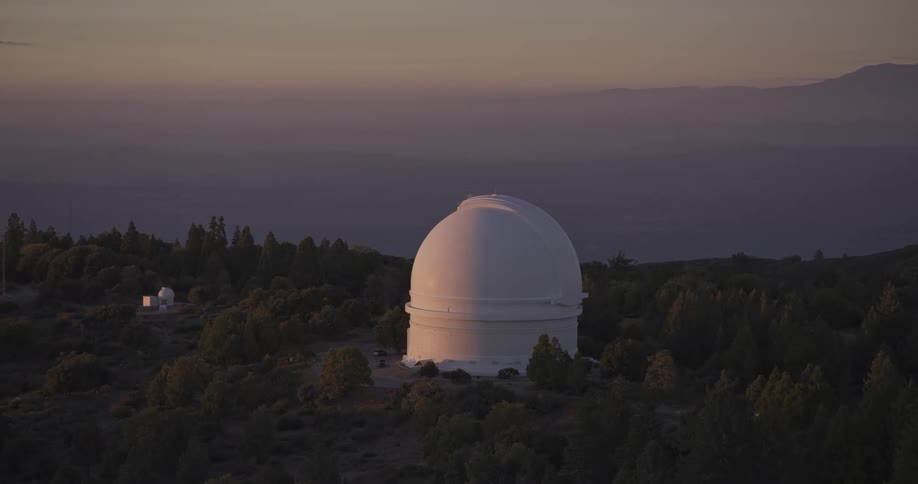 The video is an aerial or highangle shot of a large white astronomical observatory dome situated on a forested mountain ridge the scene is set at twilight or dawn with hazy layers of distant mountains visible against the pale sky in the backgro