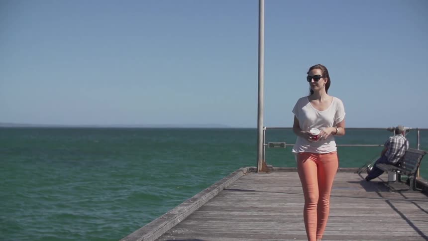 A young woman walks along a dock while holding a coffee with both hands