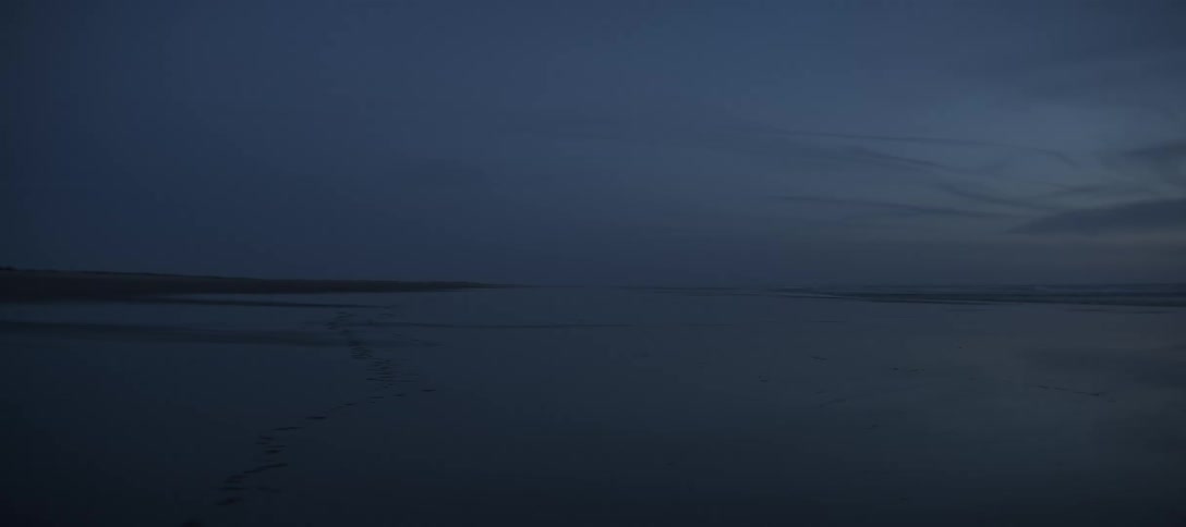 The video shows an extremely wide shot of a shoreline at twilight or night the sky is a dark deep blue matching the water and the flat land of the beach or mudflats in the foreground