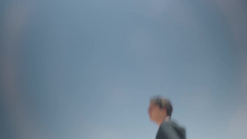 A blurred outoffocus view of a man in a dark shirt standing against a clear light blue sky with bright lens flares from the sun