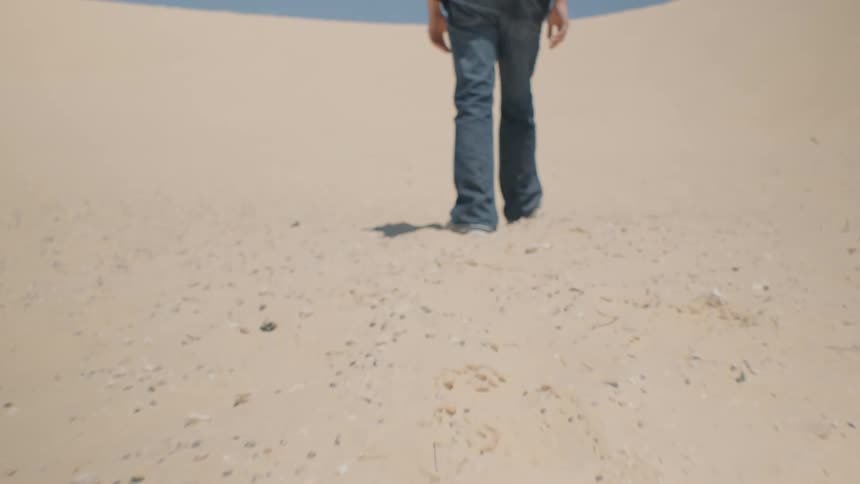 The video clip is a lowangle shot focused on a persons legs and feet walking across a bright sandy or dusty patch of ground the person is wearing dark jeans and lightcolored shoes kicking up a small amount of dust with each step