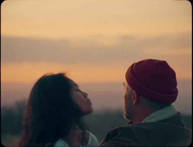 The video opens with a closeup shot of a woman smiling while cuddled into a mans chest and armsit then cuts to a twoshot of the couple turning to face each other and sharing a loving glance against a soft orange sunset backdrop