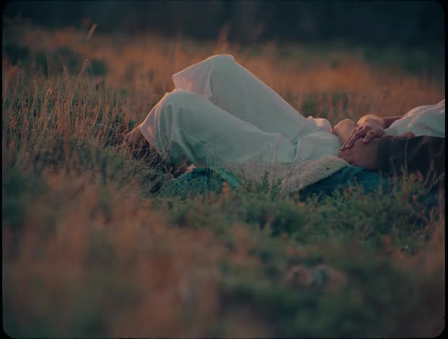 A closeup shot focuses on the lower torso and legs of a person wearing white pants lying down in a field of tall grass with their knees slightly bent remaining still in an outdoor dim light setting