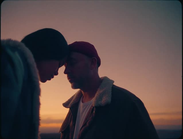 The video shows two people in near silhouette facing each other foreheads touching against a vast orange and purple sunset skythey appear to be sharing a tender intimate moment possibly for comfort or connection dressed in coldwea