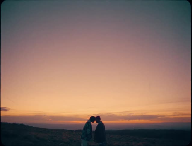 The silhouettes of two people standing close together on a hilltop against a vast sky that transitions from pink to orange near the horizon at sunset