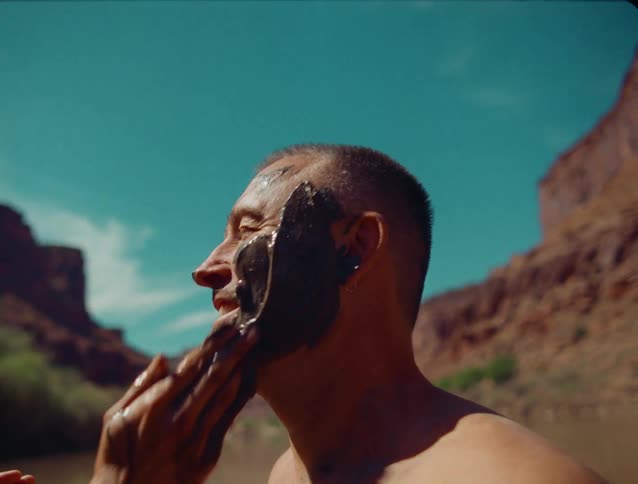 A close view shows a woman in a black top applying dark mud to a mans face for body paint followed by the man submerging and emerging from the brown silty river water against a desert canyon backdrop