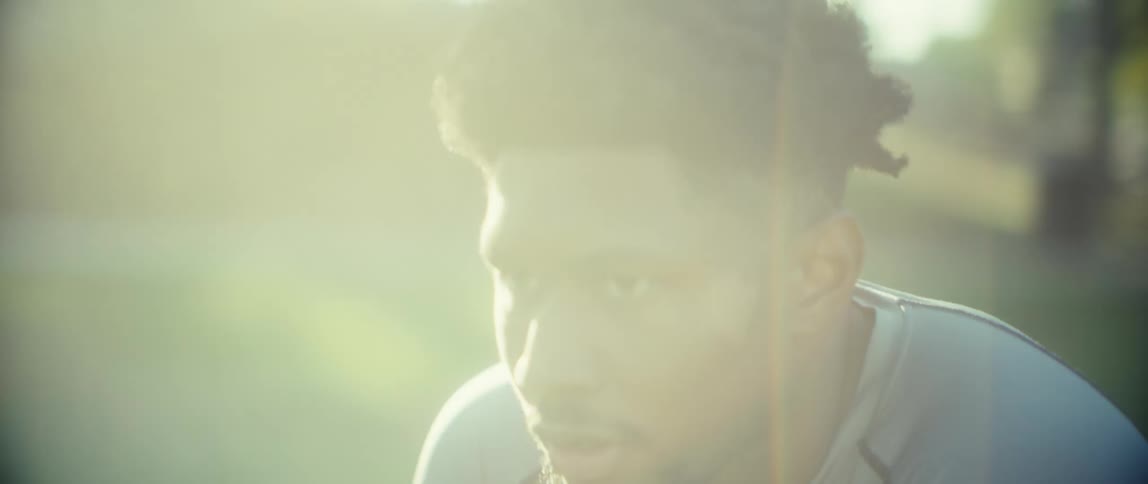 A close view shows a sweaty man with an afro hairstyle and a beard leaning over after exercise with a focused expression highlighted by bright sunlight and lens flares