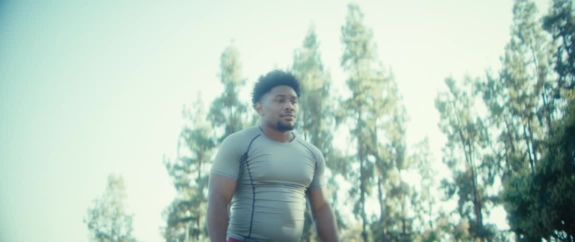 A muscular man in a tight gray athletic shirt and dark red shorts starts running or exercising outdoors under a bright sky with tall trees and sunlight flaring into the camera
