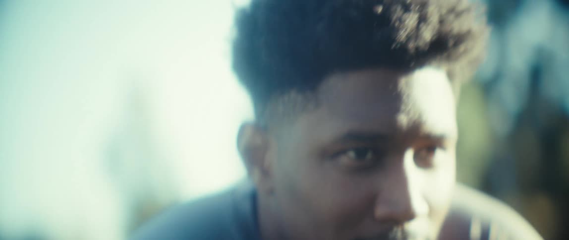 Young black man squatting on a grassy area surrounded by trees outdoors during day time