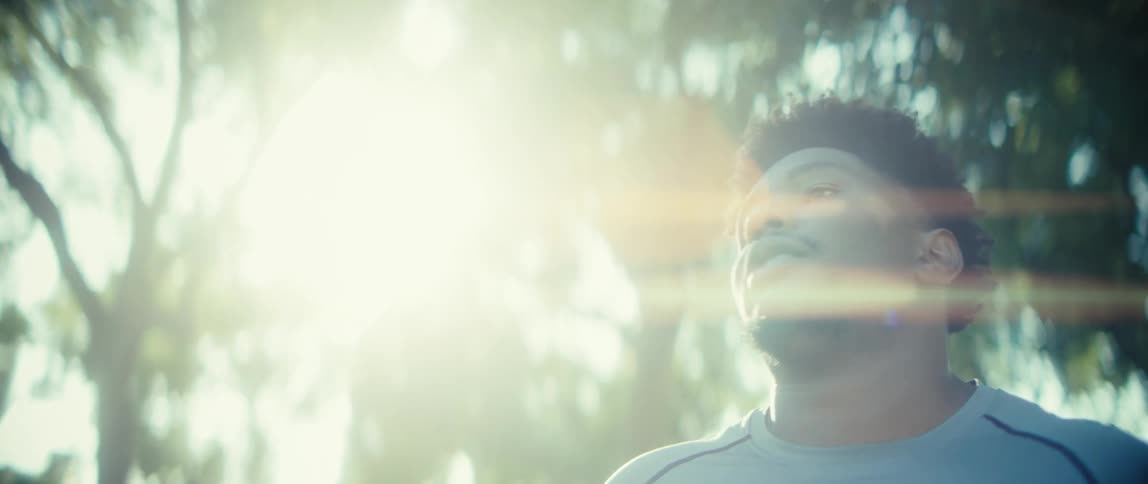 A black male on grass near trees stretches forward then places his hands behind his head appearing tired