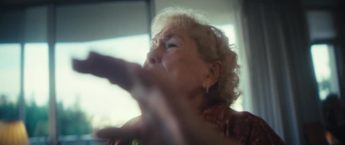 A happy older woman with blonde hair wearing a green shirt and red patterned blouse smiles and gestures excitedly while dancing energetically in a brightly lit living room