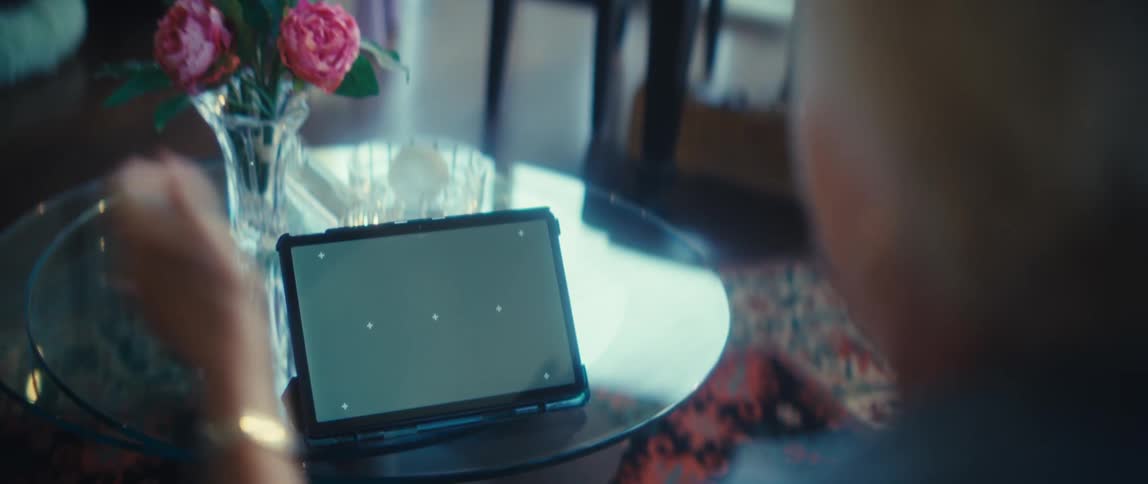 An elderly woman sits at a table having a video call on a tablet with a green screen