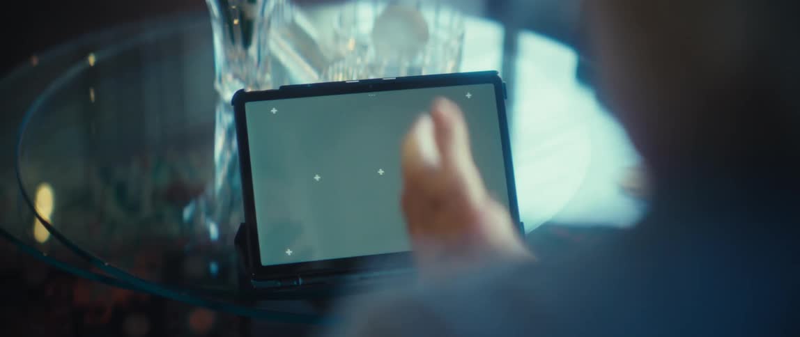 A tablet with a gray placeholder screen sits on a glass table as blurred hands clap nearby with crystal glassware in the background