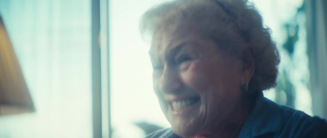 An elderly woman with white curly hair whose mouth is open wide as she displays an intense expression of powerful excitement while raising and lowering her hands in a brightly backlit indoor space