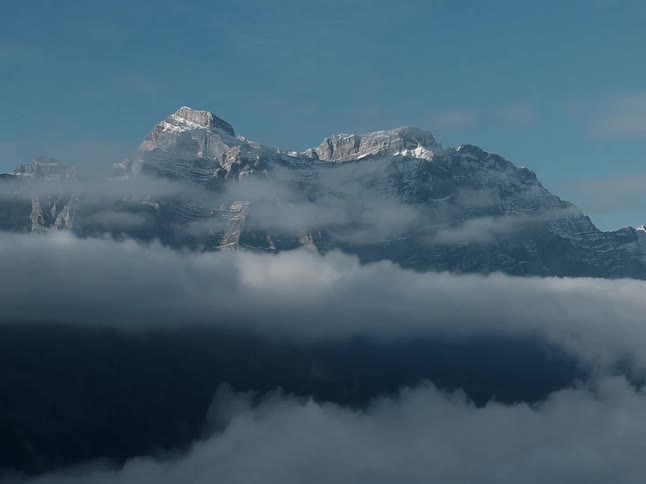 A snowdusted mountain ridge rises above a band of drifting clouds beneath a clear blue sky