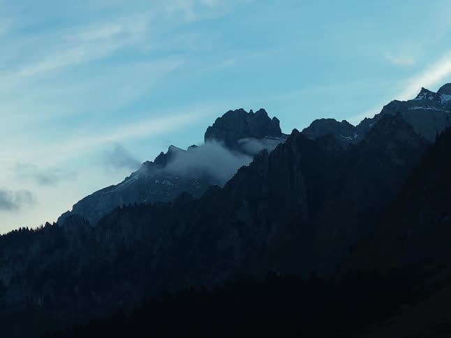 A lowangle wide view of jagged dark alpine mountain peaks shows light mist or clouds drifting across the upper slopes against a pale blue sky