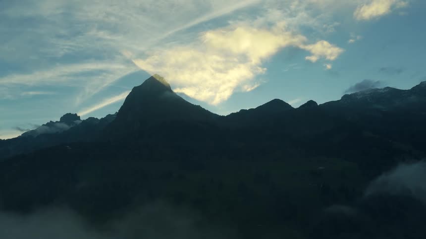 The dark silhouette of a jagged mountain range with a prominent peak centrally framed where a patch of bright sunlight illuminates a cloud just above and behind the highest point with low clouds