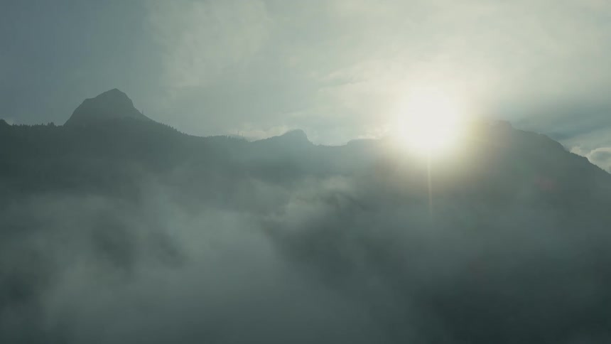 A breathtaking wide shot captures dark forested mountain peaks partially obscured by dense fog and clouds with bright sunlight and a distinct lens flare emerging over the ridge