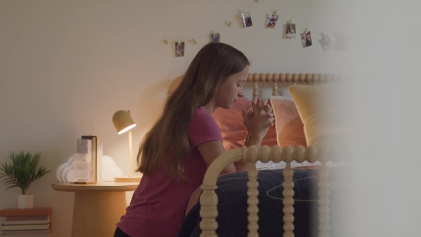 A young female teenager praying while kneeling beside her bed in her bedroom during daytime