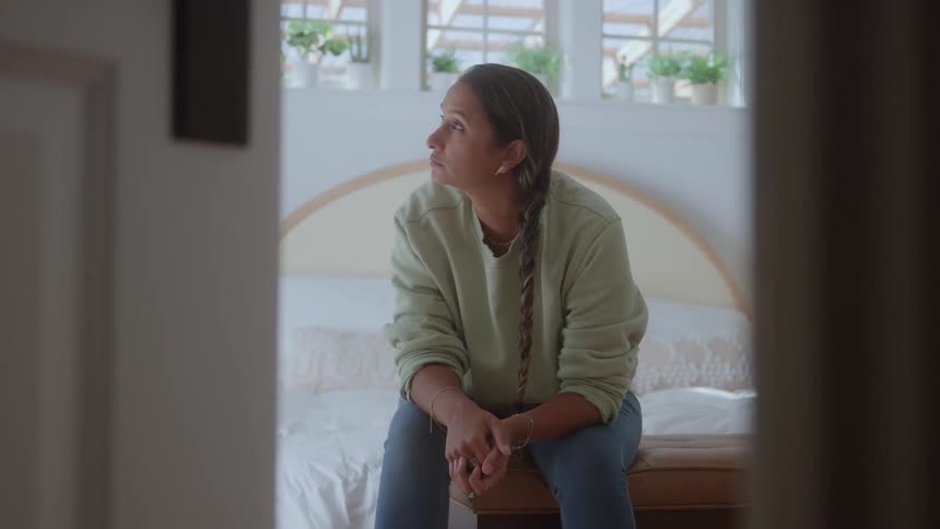 Woman in a bedroom sitting down on a bed with a contemplative face expression indoors during day time
