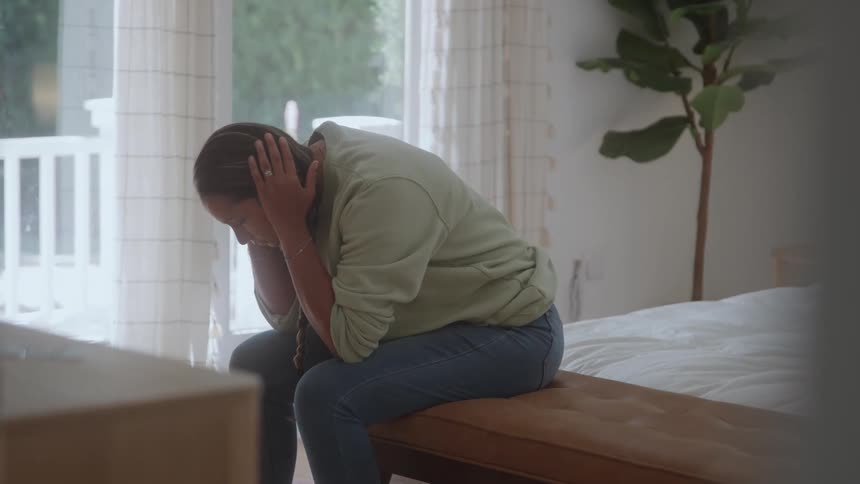 A woman sitting on a bed who is worried while holding her head in her hands