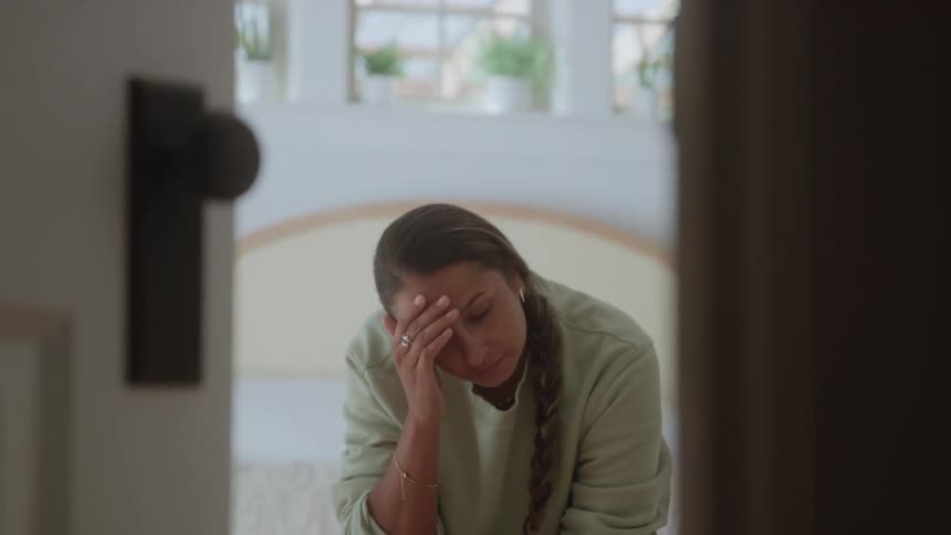 A woman with a long braid sits by a bed with her hand to her forehead looking stressed or fatigued