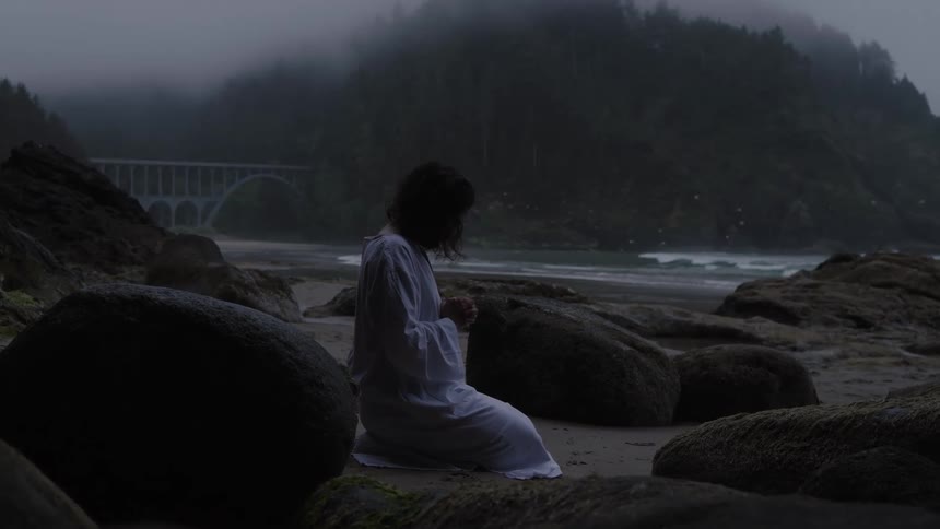 The video clip is a mediumlong shot of a person in a white robe kneeling on a dark rocky beach facing the water a large arched bridge can be seen dimly in the misty background above the beach