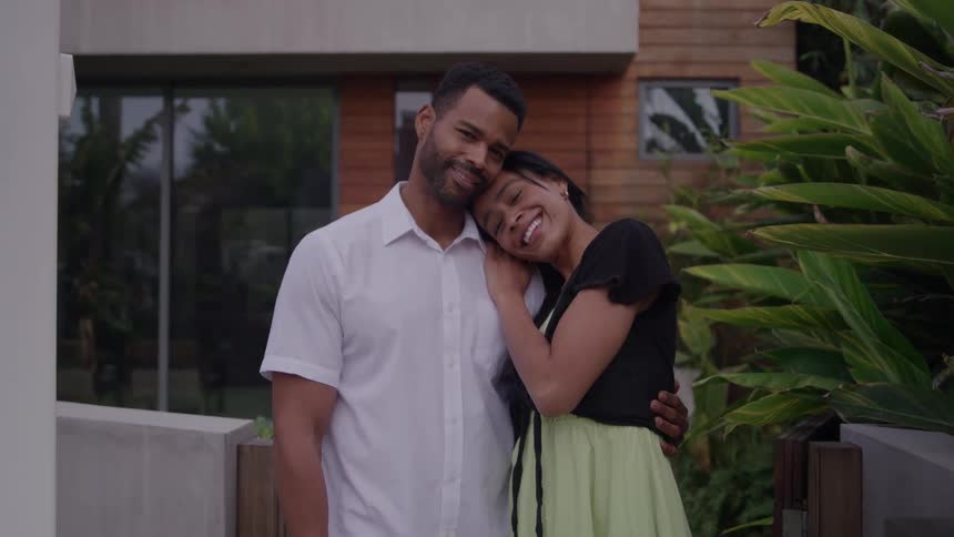 A medium view of a loving couple shows a man in a white collared shirt embracing a smiling woman in a black and light green dress outside a modern wooden house surrounded by tropical plants