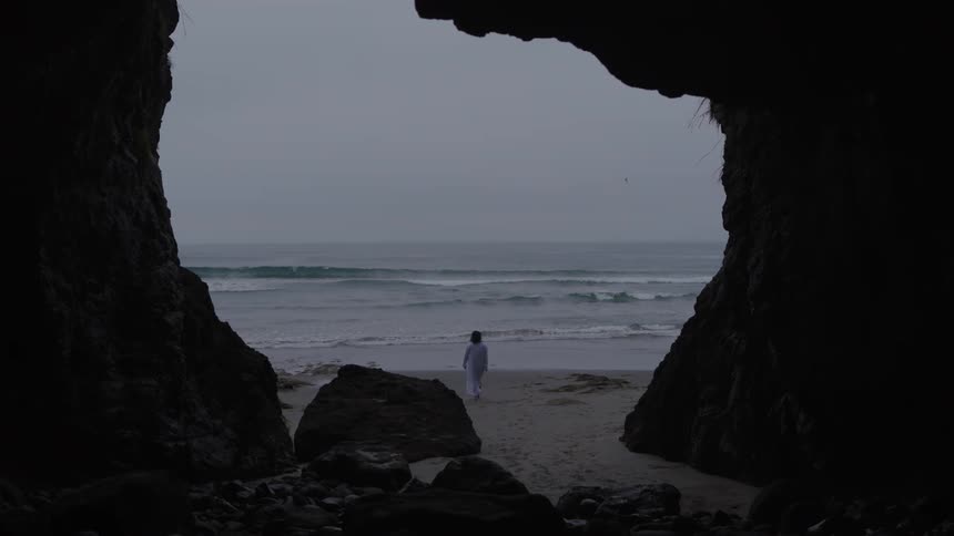 A woman wearing white clothes is coming out of a dark cave towards a beach