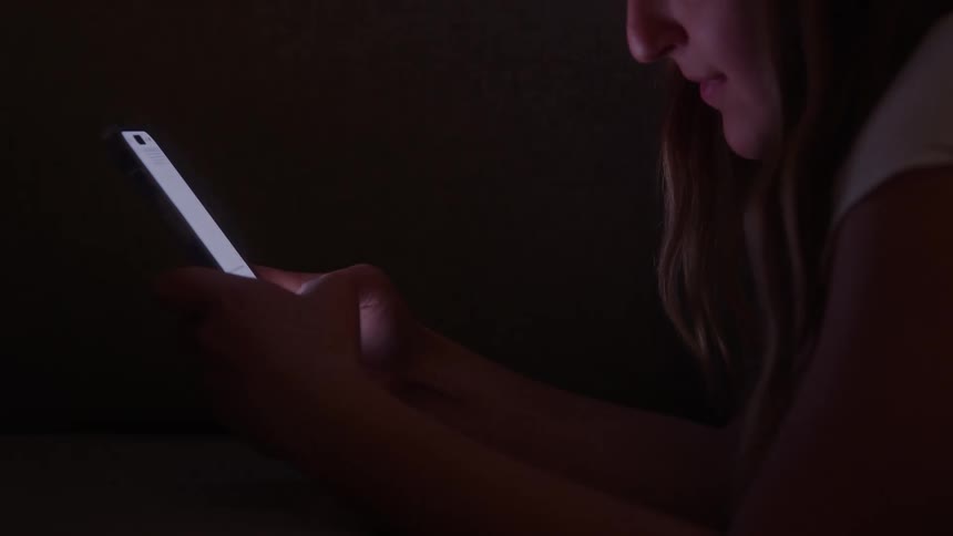 The hands and lower face of a young woman holding and typing on a smartphone in the darkness with the bright light of the screen illuminating their face and hands