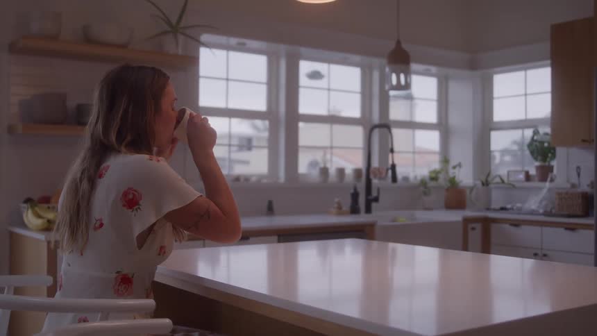 The video clip shows an indoor mediumwide shot of a bright kitchen with white countertops and whiteframed windows a woman with long hair wearing a lightcolored dress is seated at the counter holding a mug and looking out the window