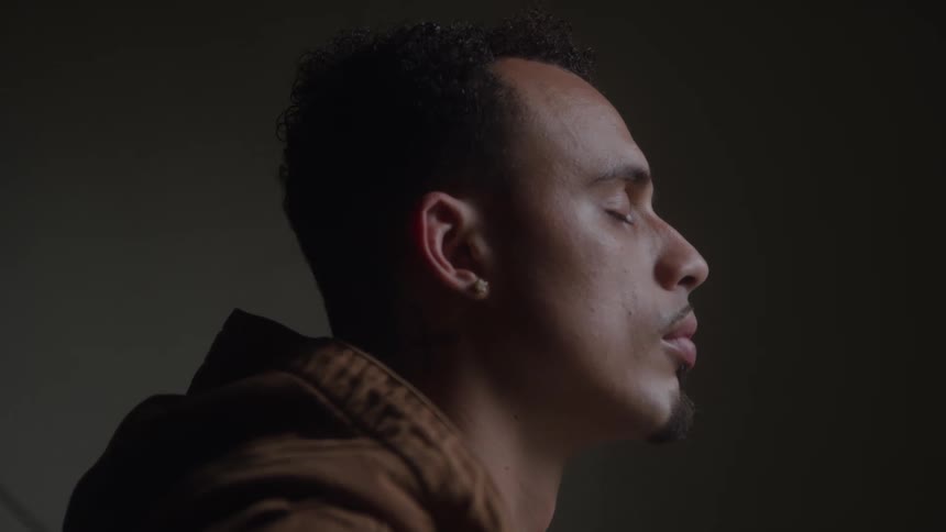 A closeup shot of a mans side profile shows him inhaling deeply and closing his eyes while he is in a dimly lit indoor space