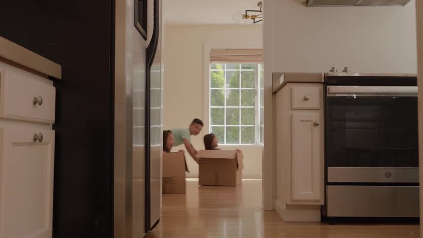 In a new house a parent pushes a child in a moving box across a hardwood floor quickly followed by another child