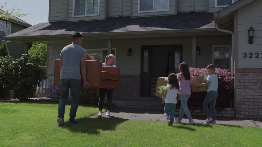 A family of five including a man and woman carrying a large wooden furniture piece and three children carrying boxes and greenery walks toward the entrance of a twostory light green suburban house on a sunny moving day