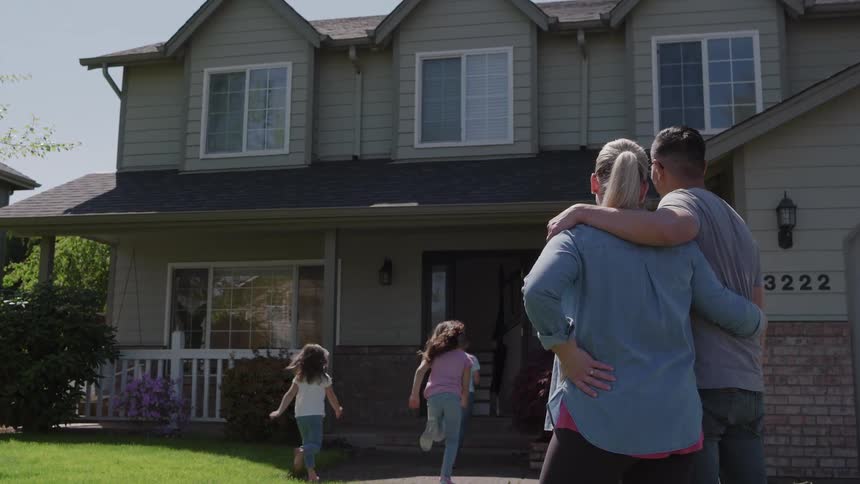 A medium view shows a man and woman hugging while admiring their new green suburban house from the lawn with two blurred children playing in the foreground
