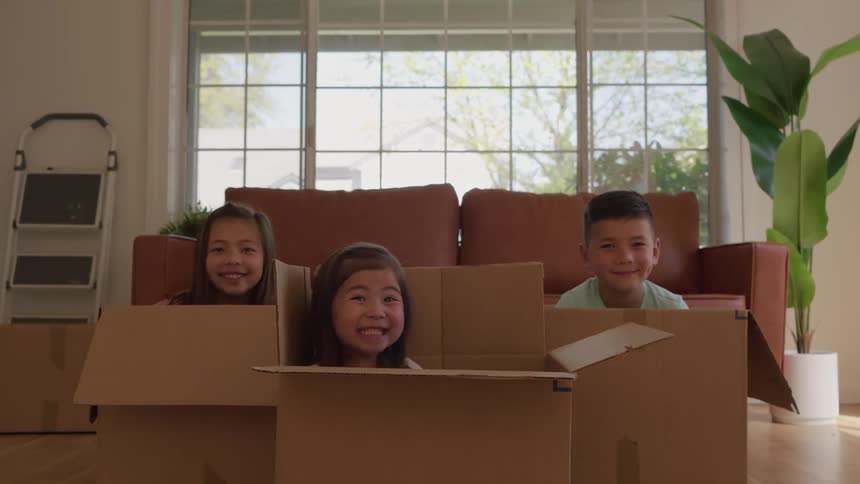 Three children two girls and a boy popping out of large cardboard moving boxes in a brightly lit room with a couch and large window behind them smiling and moving their heads in a playful manner