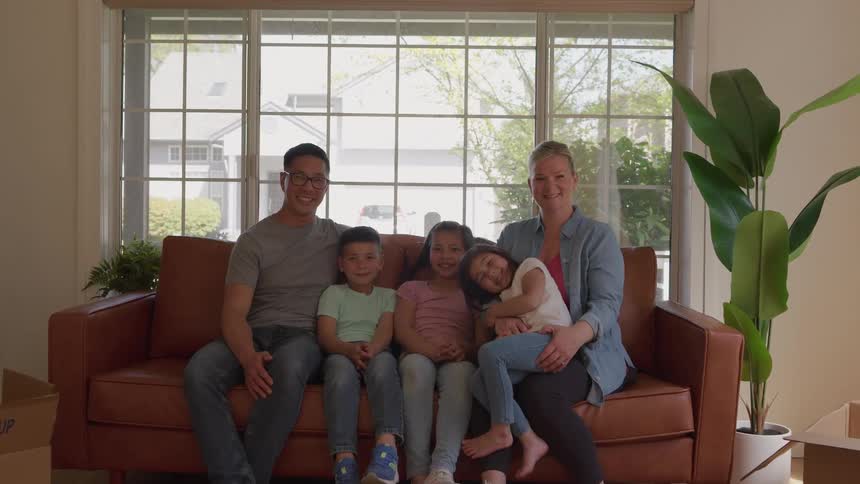 The video is a brightly lit mediumwide shot of a diverse family of four sitting on a brown leather couch in front of a large windowthe parents and their two young children are smiling directly at the camera in a room surrounded by moving box