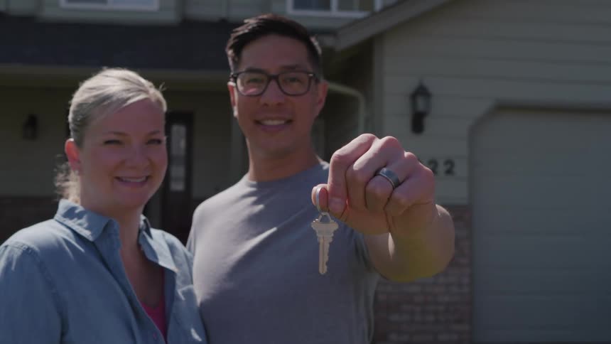 A happy couple stands smiling in front of their new light green house with the man wearing glasses and a gray shirt extending his hand toward the camera to show off a new house key