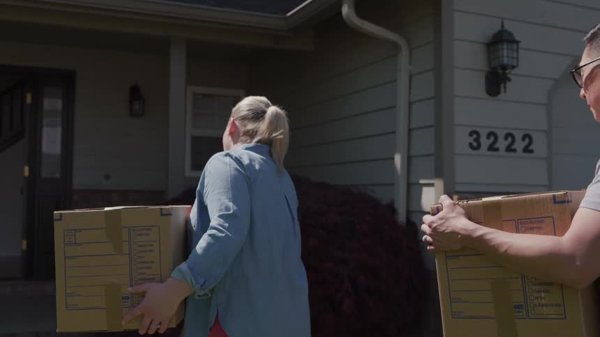 The video is a medium shot of a diverse couple a man and a woman carrying cardboard moving boxes toward the front entrance of a suburban homethe house number 3222 is visible above the garage suggesting they are moving into their new