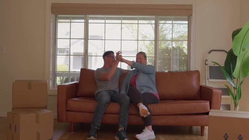 A man and a woman stand sit down on a leather sofa raise their hands and perform a hand slap gesture then sit holding hands in a living room with boxes and a large window