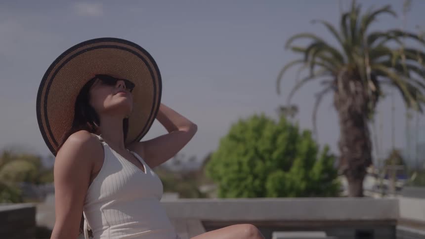 The video is a mediumcloseup shot of a woman in a white swimsuit and a widebrimmed straw hatshe is sunning herself outdoors in a bright tropicallooking setting with palm trees visible in the background