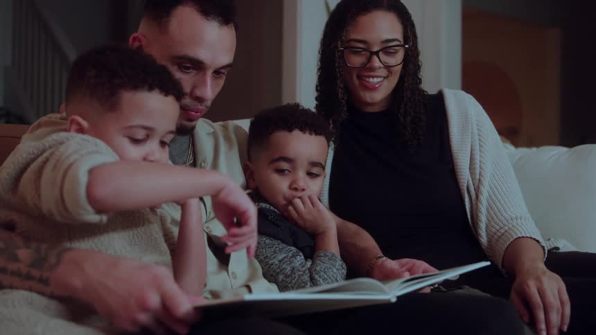 The video shows a diverse family of foura man and a woman sitting with two young boysreading a book together on a couchthey are in a cozy dimly lit living room sharing a quiet intimate moment as the mother smiles at the camera