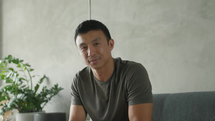 The video is a mediumcloseup shot of a man sitting indoors turning his head to smile warmly and naturally at the camerahe is casually dressed and seated on a couch in a bright room with modern minimalist decor and plants in the backgrou