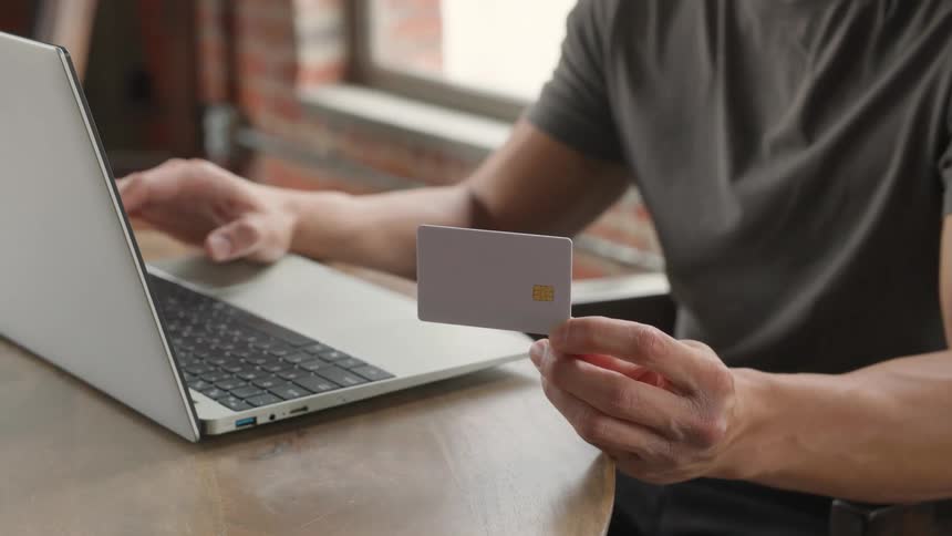 A close indoor view shows a man in a gray tshirt holding a blank gray credit card while typing payment details onto a silver laptop keyboard