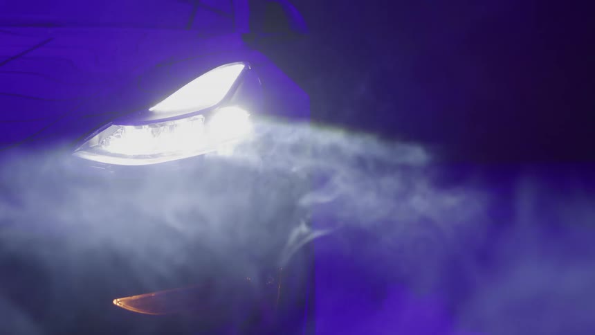 The bright led headlight of a sports car illuminates drifting smoke against a dramatic purple backdrop