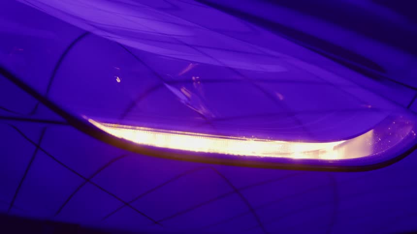 The video is a stylized closeup shot of a cars headlightrunning lightthe light emits a bright warm orangeyellow glow against the purpleblue frame of the car creating a highcontrast and dramatic effect