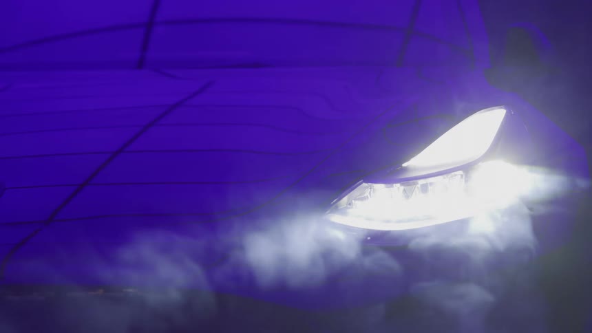 A close abstract view shows the bright white headlight of a car with visible contour lines illuminating plumes of white fog or mist against a dark purple background