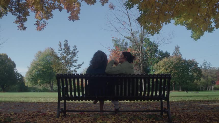 Two people are hugging each other while sitting on a public bench looking out at a lawn with trees