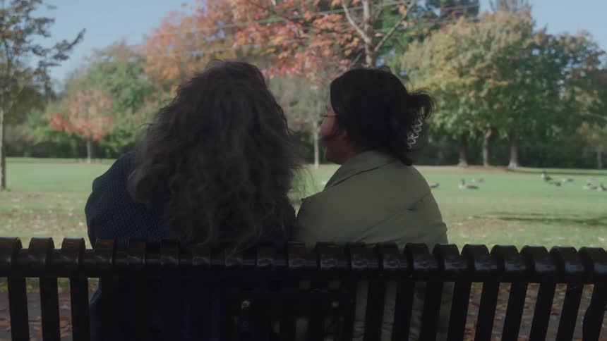 The video shows two people seated sidebyside on a park bench viewed from behind looking out at a leafy green and red park landscapethe setting suggests a quiet fall day shared between the two individuals in an outdoor space