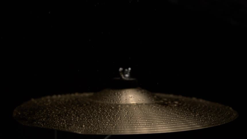 A close dramatic slowmotion shot shows a wet cymbal vibrating and shaking off thousands of small water droplets after being hit by a drumstick against a black background